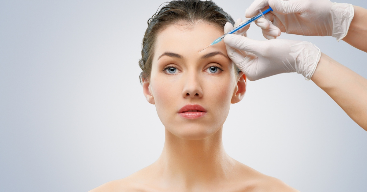 A woman with clear skin and blue eyes looks straight ahead while a medical professional wearing gloves prepares to inject a syringe into her forehead.