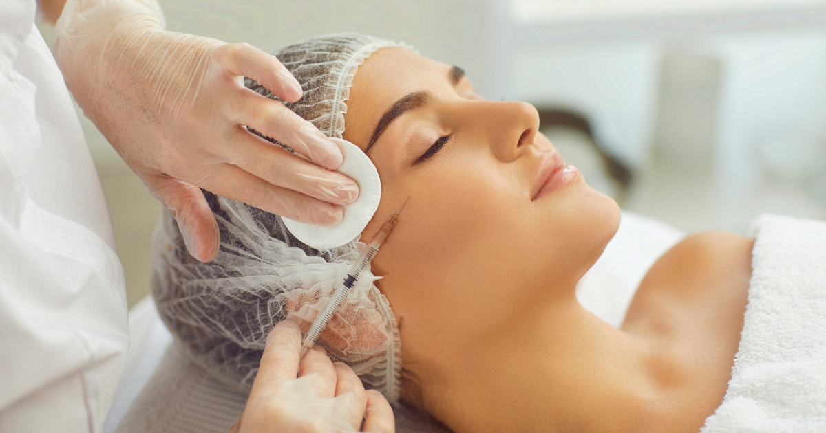 A woman lying down with her eyes closed receives a cosmetic injection near the outer corner of her eye. She is wearing a disposable hair cap while a professional in gloves administers the treatment and holds a cotton pad to her skin.