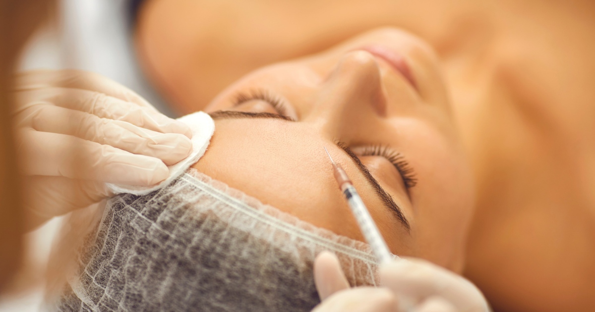 A close-up of a woman receiving a cosmetic injection on her forehead. She is lying down with her eyes closed, wearing a disposable hair cap, while a professional wearing gloves administers the injection and holds a cotton pad on her skin.